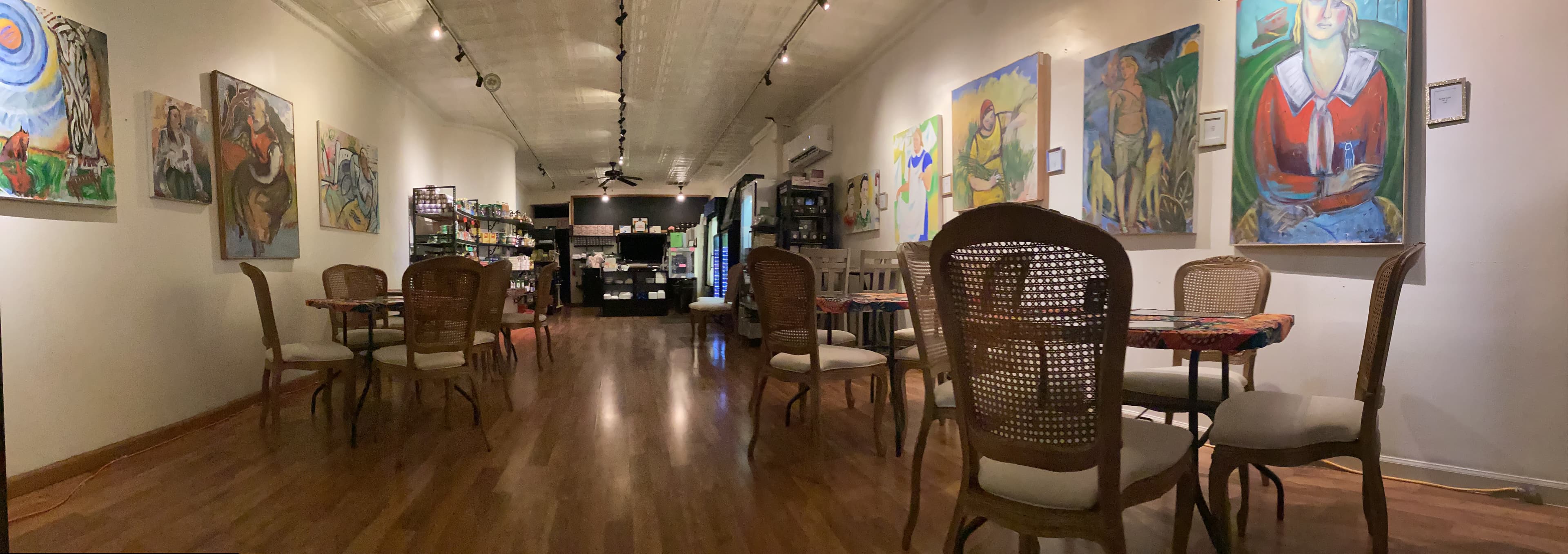 Cozy cafe interior with wooden floors, woven chairs, and colorful paintings lining the walls.